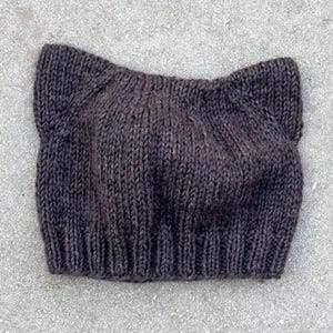 Puede incluir: Un gorro de punto marrón oscuro con orejas de gato. El gorro está hecho de un hilo grueso y texturizado y tiene un borde acanalado. Las orejas de gato son puntiagudas y sobresalen de la parte superior del gorro. El gorro es de color sólido.