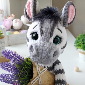 Könnte beinhalten: Ein handgehäkeltes Zebra-Plüschtier mit einem Lavendelstrauß. Das Zebra hat einen weiß-grau gestreiften Körper, große blaue Augen und rosa gefütterte Ohren. Der Strauß ist in Papier gewickelt und mit einem Band gebunden.
