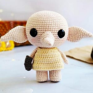 Könnte beinhalten: Handgefertigte Häkelpuppe in hellem Beige, mit großen schwarzen Augen, spitzen Ohren und einer kleinen Nase. Die Puppe trägt ein gelbes Kleid und hält einen dunkelbraunen Gegenstand. Ein skurriles, handgefertigtes Produkt.
