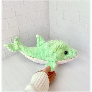May include: A handmade, light green crochet dolphin plush toy with a white belly. The dolphin has a dorsal fin, tail fins, and side fins. The toy has black safety eyes and is being held by a person.