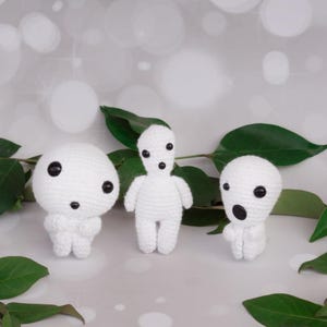 Puede incluir: Tres figuras blancas hechas a mano con ganchillo, con ojos de botón negros y rasgos sencillos. Las figuras están dispuestas con hojas verdes en el fondo. Las figuras tienen diferentes formas.