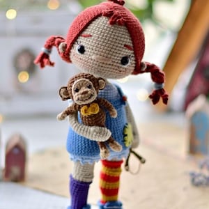 Könnte beinhalten: Gehäkelte Puppe mit roten Zöpfen, trägt ein blaues Kleid, lila Socken und blaue Schuhe. Die Puppe hält einen kleinen braunen gehäkelten Affen mit einer gelben Schleife. Die Puppe hat schwarze Augen und einen neutralen Hautton.