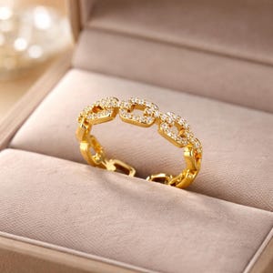Peut inclure: Une bague dorée avec un motif de chaîne, ornée de petits pierres précieuses transparentes. La bague est présentée dans un écrin à bijoux avec un intérieur de couleur neutre.