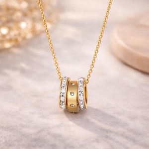 Peut inclure: Un collier doré avec trois anneaux connectés. Deux anneaux sont sertis de cristaux transparents, et l'anneau central comporte trois petits cristaux transparents. Le collier est suspendu à une fine chaîne dorée.
