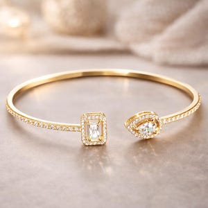 Peut inclure: Un bracelet manchette ouvert de couleur or avec deux extrémités décoratives. Une extrémité présente une pierre précieuse rectangulaire taille émeraude, et l'autre une pierre en forme de poire, toutes deux entourées de petites pierres claires. Le bracelet est posé sur un fond neutre et doux.