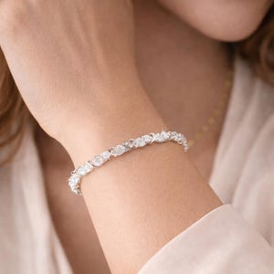 Peut inclure: Un bracelet argenté orné de nombreux pierres précieuses claires de forme ovale. Le bracelet est porté au poignet, les pierres étant rapprochées pour un éclat continu. Il s'agit d'un bijou.