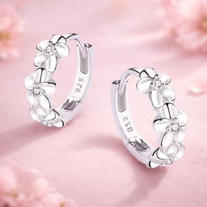 Peut inclure: Une paire de boucles d'oreilles créoles en argent. Chaque boucle présente une rangée de motifs blancs en forme de fleurs, avec une petite gemme transparente au centre. Fond rose clair.