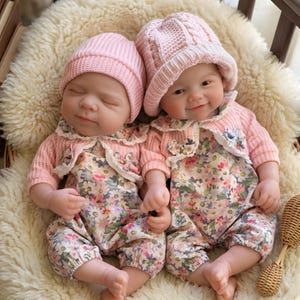 May include: Two baby dolls wearing matching pink knit hats and sweaters with floral print rompers. One doll has its eyes closed, while the other smiles. They are lying on a fluffy, white surface.