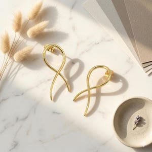 May include: Two gold-coloured, abstract ribbon-shaped earrings. The earrings are presented on a white marble surface, with a small ceramic dish and dried flowers in the background. The earrings have a post-back closure.
