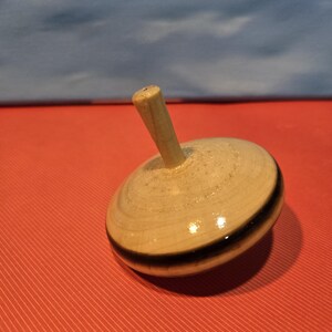 May include: A wooden spinning top with a rounded body and a handle. The top is light-colored with a black stripe around the middle. The handle is a light brown color. The top is sitting on a red surface.