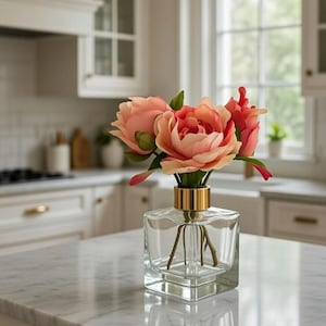 May include: A clear glass square vase with a gold rim holds several peach and red artificial flowers. The vase sits on a white marble countertop in a bright kitchen setting.