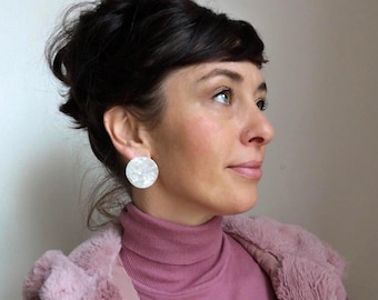 Könnte beinhalten: Eine Frau mit dunklem Haar trägt große, weiße, runde Ohrringe. Die Ohrringe haben eine strukturierte, marmorierte Optik. Sie trägt einen rosa Rollkragenpullover und eine rosa Kunstfellweste.