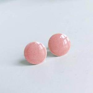 May include: Two round, pink stud earrings with a glossy finish. The earrings have a speckled texture and a simple, minimalist design. The earrings are set against a white background.
