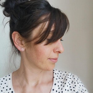 May include: A woman with dark brown hair styled in a bun and bangs. She is wearing a white top with black polka dots and a small pink stud earring. A silver nose ring is also visible.