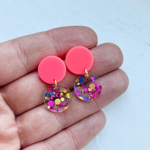 Könnte beinhalten: Ein Paar Ohrringe mit einem neonrosa Kreis und einem transparenten Kreis, der mit Glitzer gefüllt ist. Der obere Kreis ist einfarbig neonrosa. Der untere Kreis ist transparent mit rosa, blauen, gelben und lila Glitzer. Die Ohrringe sind durch eine goldfarbene Schlaufe verbunden.