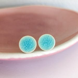 Könnte beinhalten: Ein Paar runde, türkisfarbene Keramik-Ohrstecker mit Craquelé-Effekt. Die Ohrringe liegen auf einer rosa Oberfläche. Diese Ohrringe sind handgefertigt.