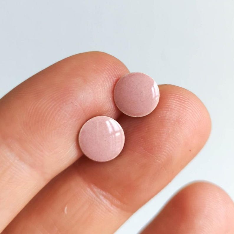 May include: Two small, round, light pink earrings with a glossy finish. The earrings are held between fingers against a white background. The earrings are a simple, minimalist design.
