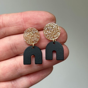 Könnte beinhalten: Ein Paar goldene Glitzer- und schwarze Bogenohrringe. Die Ohrringe sind aus Polymer Clay gefertigt und haben eine goldene Glitzer-Oberfläche. Die Bogenform ist ein modernes und trendiges Design.