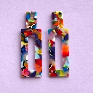 Könnte beinhalten: Ein Paar farbenfrohe rechteckige Ohrringe mit geometrischem Design. Die Ohrringe bestehen aus einem durchsichtigen Material mit einer Vielzahl von Farben, darunter Rot, Blau, Grün, Gelb und Lila.