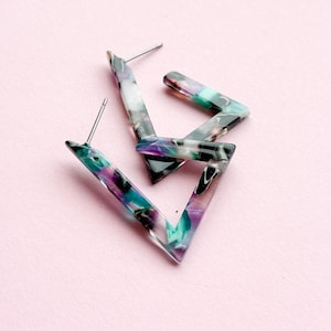 Könnte beinhalten: Geometrische Dreiecks-Ohrringe mit einem marmorierten Muster in Lila, Türkis und Weiß. Die Ohrringe haben einen silbernen Stecker und sind vor einem rosa Hintergrund platziert. Ein modernes Accessoire.