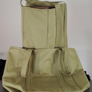 May include: Two olive-green tote bags. One bag is upright, showing the top opening and handles. The other bag is lying down, with the handles visible. Both bags are made of a sturdy fabric and have a simple, rectangular design.