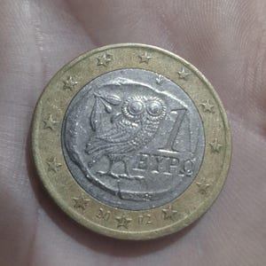 Op de afbeelding: Een euromunt van één euro met een gedetailleerd uilontwerp in het midden, omringd door een goudkleurige ring met sterren. De munt heeft het nummer "1" en het woord "EURO". Het jaartal "2002" is ook zichtbaar.