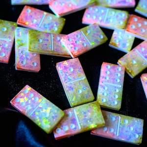 May include: A collection of colorful dominoes with a shimmering, iridescent effect. The dominoes have a rectangular shape with rounded corners and white dots. The colors include pink, yellow, and green, with a glittery finish. The dominoes are scattered on a black surface.