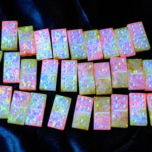 May include: A set of iridescent dominoes with a glittery, colourful appearance. The dominoes have a pink, yellow, and blue colour scheme, with white dots. The game pieces are arranged on a dark, textured surface.