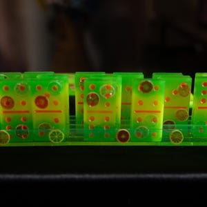Puede incluir: Un juego de dominó verde y amarillo con diseños circulares naranjas y rojos. Los dominós están dispuestos sobre un soporte verde a juego. Los dominós están hechos de un material translúcido.