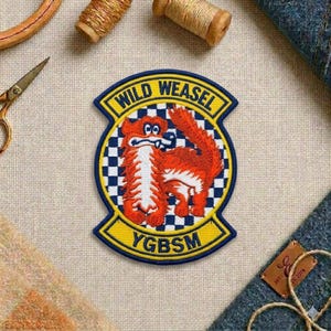 May include: Embroidered patch featuring a cartoon weasel in orange and white, set against a blue and white checkered background. The patch has a yellow border with the words "WILD WEASEL" and "YGBSM".