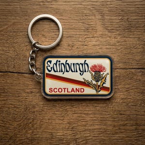 May include: A rectangular keychain featuring the text "Edinburgh SCOTLAND" with a thistle graphic. The keychain has a silver-colored ring and chain. The design is set against a cream background with red and brown stripes.