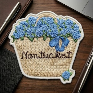 May include: An embroidered patch featuring a tan woven basket filled with blue hydrangea flowers and green leaves. A blue ribbon is tied to the handle. The word Nantucket is stitched in brown thread across the center of the basket.