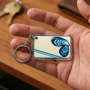 May include: A rectangular keychain with a blue and white seashell design. The keychain has a silver metal ring and chain. The background of the keychain is off-white with blue stripes.