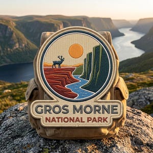 May include: A tan backpack featuring a circular embroidered patch with a landscape scene of a caribou standing on a red cliff edge, a blue river, green cliffs, and an orange sun. The text GROS MORNE NATIONAL PARK is embroidered below the circle.