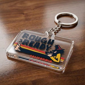 May include: A clear rectangular keychain with the words "ROUGH READY" in black and a graphic of a person in a hat. The keychain has a silver key ring and chain. The text "CALIFORNIA" is printed in red.