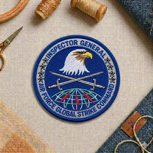 May include: A circular blue patch with white and gold text that reads "INSPECTOR GENERAL AIR FORCE GLOBAL STRIKE COMMAND". The patch features an eagle, crossed swords, and a globe design.