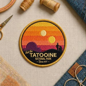 May include: A circular embroidered patch featuring a sunset landscape with two yellow suns, a purple desert structure, and two black silhouettes of figures. The text reads Visit the TATOOINE NATIONAL PARK Since 1977 in yellow embroidery.