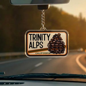 May include: A rectangular keychain with the words "TRINITY ALPS CALIFORNIA" and a brown pinecone illustration. The keychain is hanging from a silver chain, with a blurred background of a road and trees.