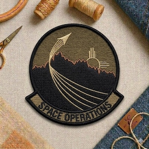 May include: Embroidered patch with a circular design featuring a stylized rocket launching from a mountain range, with the text "SPACE OPERATIONS" in a banner below. The patch has a black border and earth-tone colors.