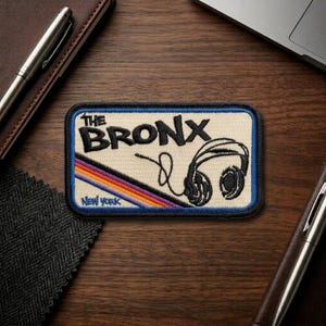 May include: Embroidered patch featuring the text "The Bronx" in black, with a rainbow design and a pair of headphones. The patch also includes the text "New York" in blue. The patch has a blue border.