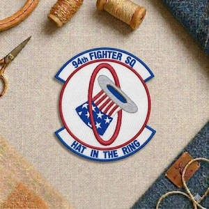 May include: Embroidered patch with the text "94th FIGHTER SQ" and "HAT IN THE RING". The patch features a design with red, white, and blue elements, including a stylized hat and stars. The patch is on a beige fabric.