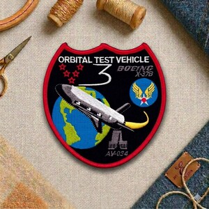 May include: Embroidered patch with a black background and a red border. The patch features a spacecraft over a blue and green globe, with the text "ORBITAL TEST VEHICLE 3 BOEING X-37B" and "AV-034".