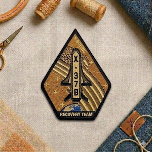 May include: A diamond-shaped patch with a black border features the text "BOEING" and "X-37B". The patch has a brown and gold color scheme with a depiction of a spacecraft and the words "RECOVERY TEAM".