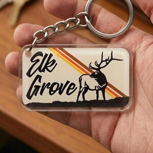 May include: A rectangular keychain with the words "Elk Grove" in black script. The background is cream-colored with diagonal stripes of orange, yellow, and brown. A black and white elk silhouette is on the right.