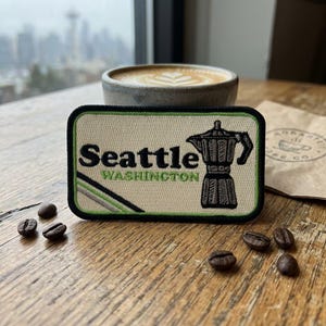 May include: A rectangular embroidered patch with a cream background and a black border. It features the black text Seattle, the green text WASHINGTON, and a black moka pot illustration. A diagonal green and grey stripe accents the bottom left corner.