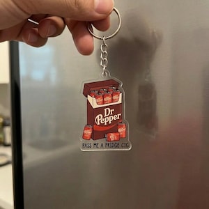May include: A novelty keychain featuring a Dr Pepper-themed design. The keychain depicts a box of Dr Pepper cans with the text "Dr Pepper" and "PASS ME A FRIDGE CIG". The keychain is attached to a silver keyring.