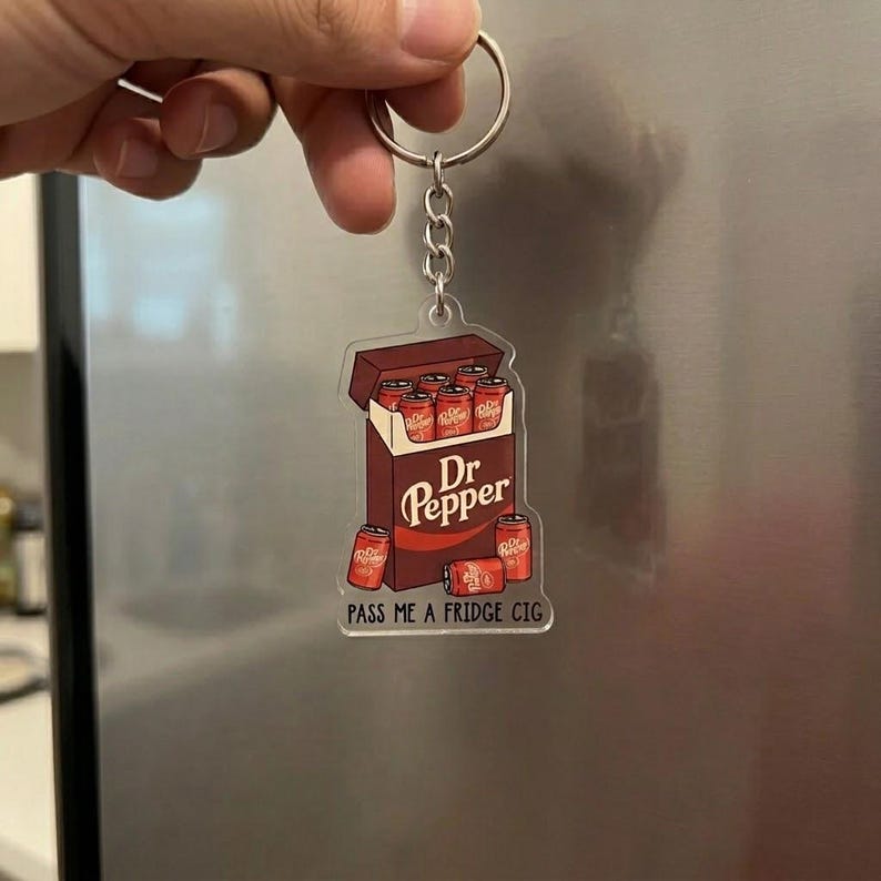 May include: A novelty keychain featuring a Dr Pepper-themed design. The keychain depicts a box of Dr Pepper cans with the text "Dr Pepper" and "PASS ME A FRIDGE CIG". The keychain is attached to a silver keyring.