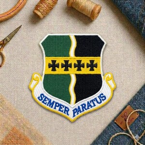 May include: Embroidered patch with a shield design. The patch features green and black sections, yellow crosses, and the words "Semper Paratus" in blue. The border is white and yellow.