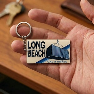 May include: A rectangular keychain with the text "LONG BEACH CALIFORNIA" and an illustration of a shipping container. The keychain has a silver ring and chain. The background is a cream color with blue stripes.