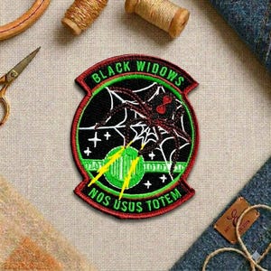 May include: Embroidered patch with a black widow spider design. The patch features a black background with a spider web and a red spider. The text "BLACK WIDOWS" and "NOS USUS TOTEM" are in green and red.